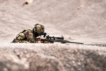 Sniper Soldier in Full Camouflage Aiming from Concealed Position in Rocky Desert Terrain –...