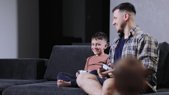 Father and his little son sit on sofa in living room playing video games together at home.