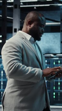 Vertical video Manager walking in server hub using tablet to find firewall misconfigurations affecting rackmounts performance. Ad,om checking systems bottlenecks leading to sluggish data transfer