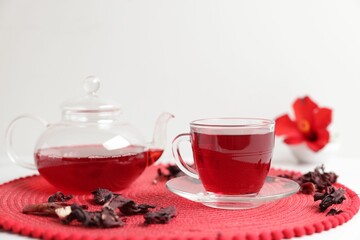 Delicious hibiscus tea in glass cup, teapot and dry roselle sepals on white table, closeup