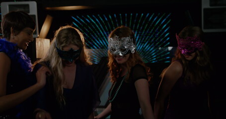 Posing four women wearing masquerade masks at dimly lit party venue, with radial neon lights