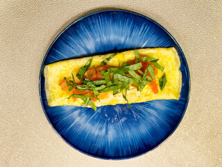 Morning breakfast meal of a large egg omelet with sliced tomato and basil on dark blue plate in overhead flat lay view