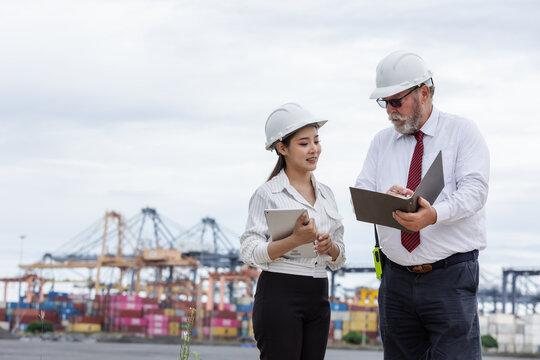 A supervisor and secretary discuss logistics operations at a busy shipping port. Both wear safety helmets and hold documents, coordinating work amid cranes and stacked cargo containers.