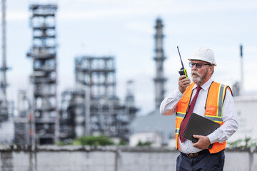 A senior engineer in a safety vest and helmet uses a walkie-talkie while holding a clipboard at an oil refinery. Industrial towers in the background reflect a high-level site inspection.