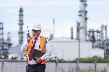 A senior engineer in a safety vest and helmet uses a walkie-talkie while holding a clipboard at an oil refinery. Industrial towers in the background reflect a high-level site inspection.