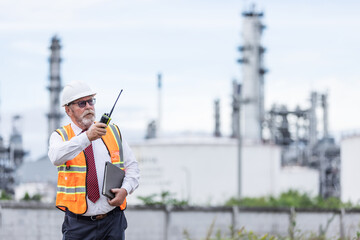 A senior engineer in a safety vest and helmet uses a walkie-talkie while holding a clipboard at an oil refinery. Industrial towers in the background reflect a high-level site inspection.