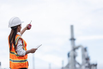 A female engineer in a safety vest and helmet communicates via walkie-talkie while holding a tablet. She observes operations at an industrial oil refinery site in the background.