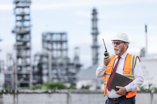 Senior engineer at an oil refinery wearing safety gear, using a walkie-talkie and holding documents while overseeing industrial operations with refinery towers in the background.