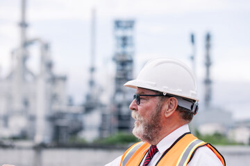 A senior male engineer in a white helmet and safety vest observes an oil refinery site. Industrial...