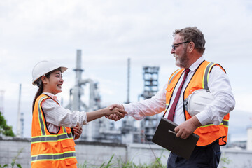 Two engineers shake hands at an oil refinery site, symbolizing successful collaboration or agreement. Both wear safety gear, with refinery structures visible in the background.