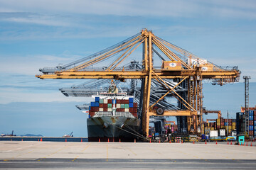 A large cargo ship is docked at a commercial port with towering cranes loading and unloading...