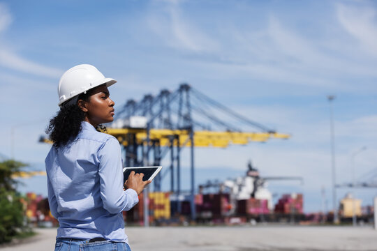 A female engineer in a safety helmet uses a digital tablet while monitoring container logistics at a shipping port. Cranes and trucks in the background show active freight operations.