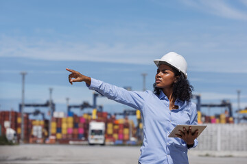 A female manager wearing a helmet and holding a tablet points forward at a busy shipping port. Surrounded by containers and trucks, she oversees logistics and directs port operations.