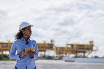 A female engineer wearing a safety helmet holds a tablet while inspecting the construction site of oil rigs. Heavy structures in the background highlight offshore industry development.