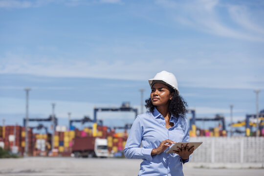 A female engineer at a shipping port uses a tablet and talks on the phone while wearing a safety helmet. She applies technology for real-time communication and logistics management.