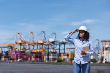 A female engineer at a shipping port shades her eyes while observing port operations. Holding a tablet, she monitors cargo logistics with cranes and colorful containers in the background.