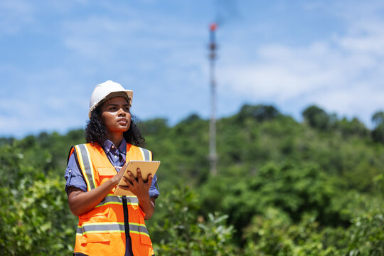 A female environmental engineer in safety gear takes notes outdoors near a forested area with a visible industrial chimney, indicating monitoring or assessment of air pollution impact.