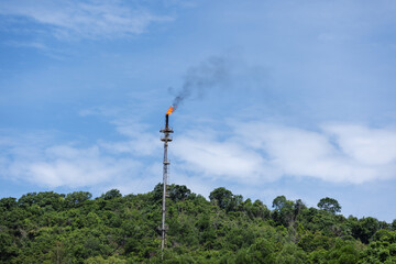 Obraz premium A gas flare emits smoke and flames from a tall industrial stack situated near a forested area, symbolizing air pollution and environmental impact from oil or gas processing activities.