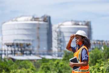 An environmental engineer conducts a site survey near large industrial storage tanks. Wearing safety gear and holding a tablet, she evaluates environmental impact and infrastructure integrity.