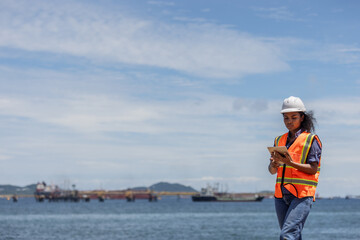 A female engineer conducts a coastal survey near the shore, holding a tablet and wearing safety...