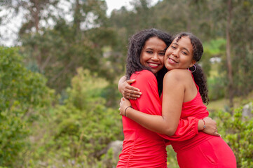 Two smiling sisters hugging each other, wearing red dresses in nature