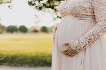 Close-up of pregnant woman in pink lace dress gently cradling baby bump with golden field and bokeh...