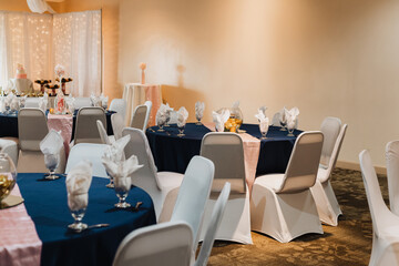 Spacious wedding reception room with round tables draped in navy linens and blush runners, white chair covers, water-glass napkins, inverted glass bowl centerpieces with gold shells and candles, and f