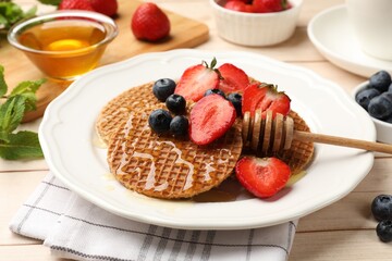 Tasty Dutch waffles (stroopwafels) with honey, strawberries and blueberries on light wooden table, closeup
