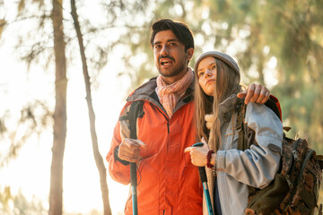 Romantic Couple Hiking in the Forest During Golden Hour. Embracing While Watching Sunset Light Through Trees, Wearing Warm Jackets and Backpacks, Feeling Connected and Adventurous in Nature