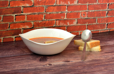 Solyanka, a tasty Russian soup with a lemon wedge in a deep ceramic bowl on a wooden table, a tablespoon and two pieces of wheat bread nearby.