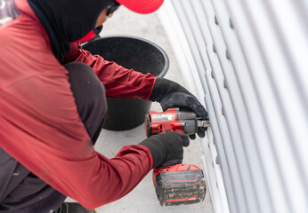 The worker is using a cordless impact wrench to tighten bolts on the metal sheet tank wall .