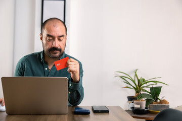 A bald Latino man at home making an online purchase on his laptop with his red credit card....