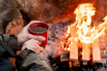 Camping lifestyle concept. Hands holding red enamel mug near a campfire.