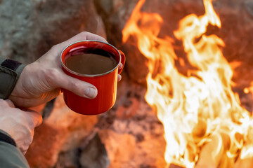Camping lifestyle concept. Hands holding red enamel mug near a campfire.