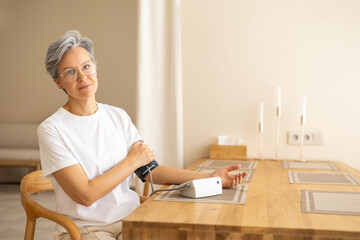 Senior woman checking blood pressure with monitor at home, sitting calmly at wooden table indoors