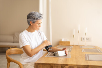 Mature woman measuring blood pressure at home using monitor while sitting at table in daylight