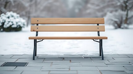 A lone wooden park bench sits on a tiled path, surrounded by a serene, snow-covered landscape with blurred trees in the background under an overcast sky.