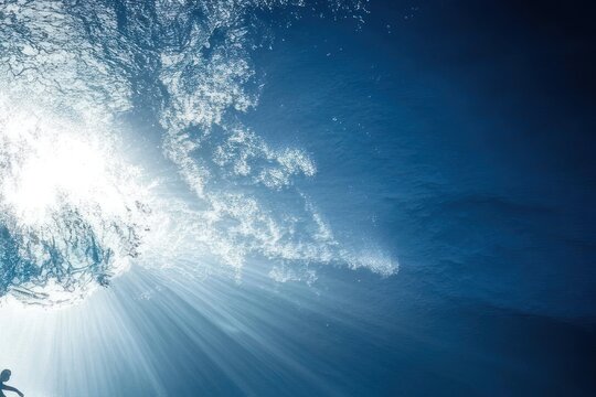 Sunlight streams through the surface of the ocean, creating a dramatic underwater scene. Silhouette of a diver or swimmer is visible. Vast, deep blue waters with white/grey waves and foam