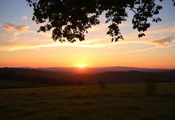 Fototapeta premium Golden sunset hues paint the sky above a tranquil meadow, viewed from beneath a leafy tree, distant hills silhouetted, horizon, dusk