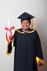 Young man wearing a cap and gown proudly shows his diploma, satisfied to finish and graduate from...