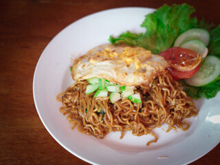 A dish of Indonesian noodles topped with a perfectly fried egg. Served on a clean white plate, this dish is complemented with fresh cucumber, lettuce, and tomato.