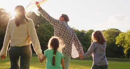 Happy family running and having fun in summer park enjoying sunny day in nature flying kite together. Mother, father and two children running on green grass outdoors at sunset. 4k video. Slow motion. - Powered by Adobe