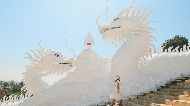 Woman tourist wear stylish white dress sightseeing explores iconic twin white dragons on temple stairs at Wat Huay Pla Kang in Chiang Rai, Thailand under clear blue sky. Famous travel phot spot