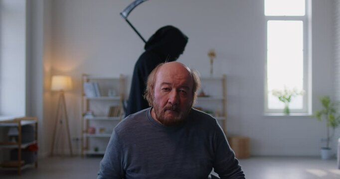 Sad senior elderly man sitting in living room at home with person in grim reaper death costume holding scythe walking behind him. Old male person haunted by death. 4k video. Slow motion video.