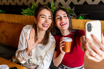 Two young women are making a video call with a smartphone, holding takeaway coffee and waving at the camera while sitting in a coffee shop