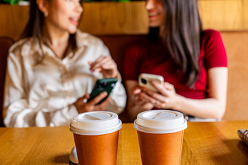 Two takeaway coffee cups sit on a wooden table in the foreground, while two blurred women use smartphones in a cafe in the background