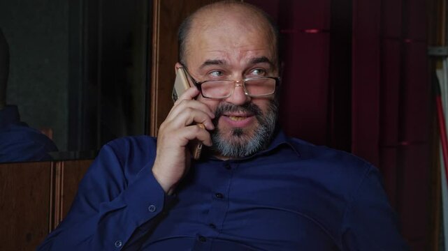 An engaging and potentially surprised or curious mood is captured as a bald man with a beard and glasses talks on a smartphone, looking off-camera with an expressive gaze