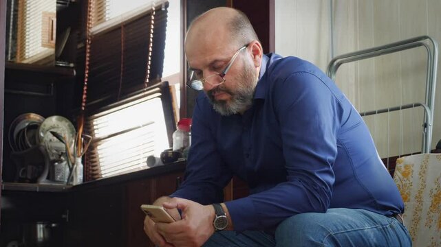 A focused and contemplative mood is captured as a bald man with a beard and glasses sits, intently looking down at his smartphone. This shot is ideal for depicting digital engagement, online research