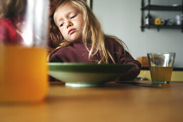 Little girl feeling sad at the kitchen table with mother nearby. childhood feelings, family...
