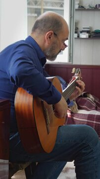 A focused and artistic mood is captured as a bald man with a beard and glasses sits and plays an acoustic guitar, his hands visible on the instrument. This shot is ideal for depicting musical talent